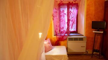 Una stanza piccola con un letto, una finestra con tende floreali, un condizionatore d'aria e una TV su un mobile.