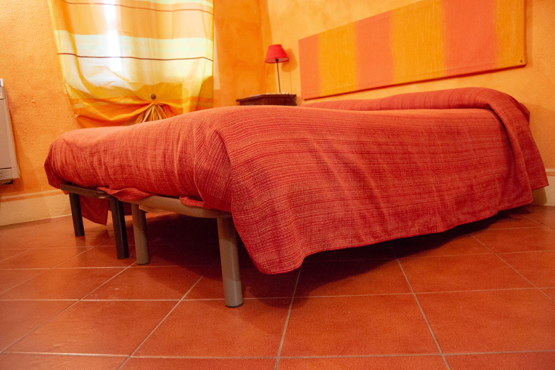 Una cama individual con una colcha roja en una habitación con paredes anaranjadas y suelo de baldosas.