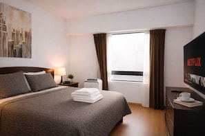 Une chambre d’hôtel avec un lit, un téléviseur diffusant Netflix, une fenêtre avec des rideaux, une table de chevet avec une lampe et un tableau accroché au mur.
