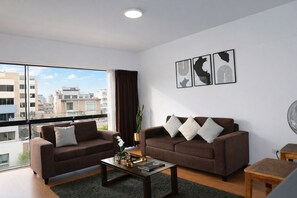Un salon moderne avec deux canapés, une table basse et une vue sur les immeubles à travers de grandes fenêtres.