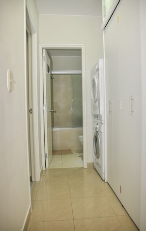 Un cuarto de lavado con lavadora y secadora, un box de ducha de vidrio, y una puerta que da a un baño.