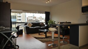 Ein modernes Wohnzimmer mit Sofa, Couchtisch und einem Glastisch für Essbereich. Es gibt eine Küchenzeile mit Mikrowelle und einer kleinen Kochinsel. Große Fenster bieten Blick auf die Gebäude draußen.