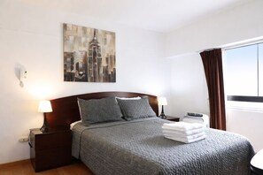 Habitación de hotel con cama, dos lámparas de noche, una mesita de noche, un cuadro de un paisaje urbano, una ventana con cortinas y una pila de toallas.