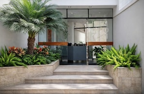 Una entrada moderna a un edificio con una palmera, helechos y un banco.