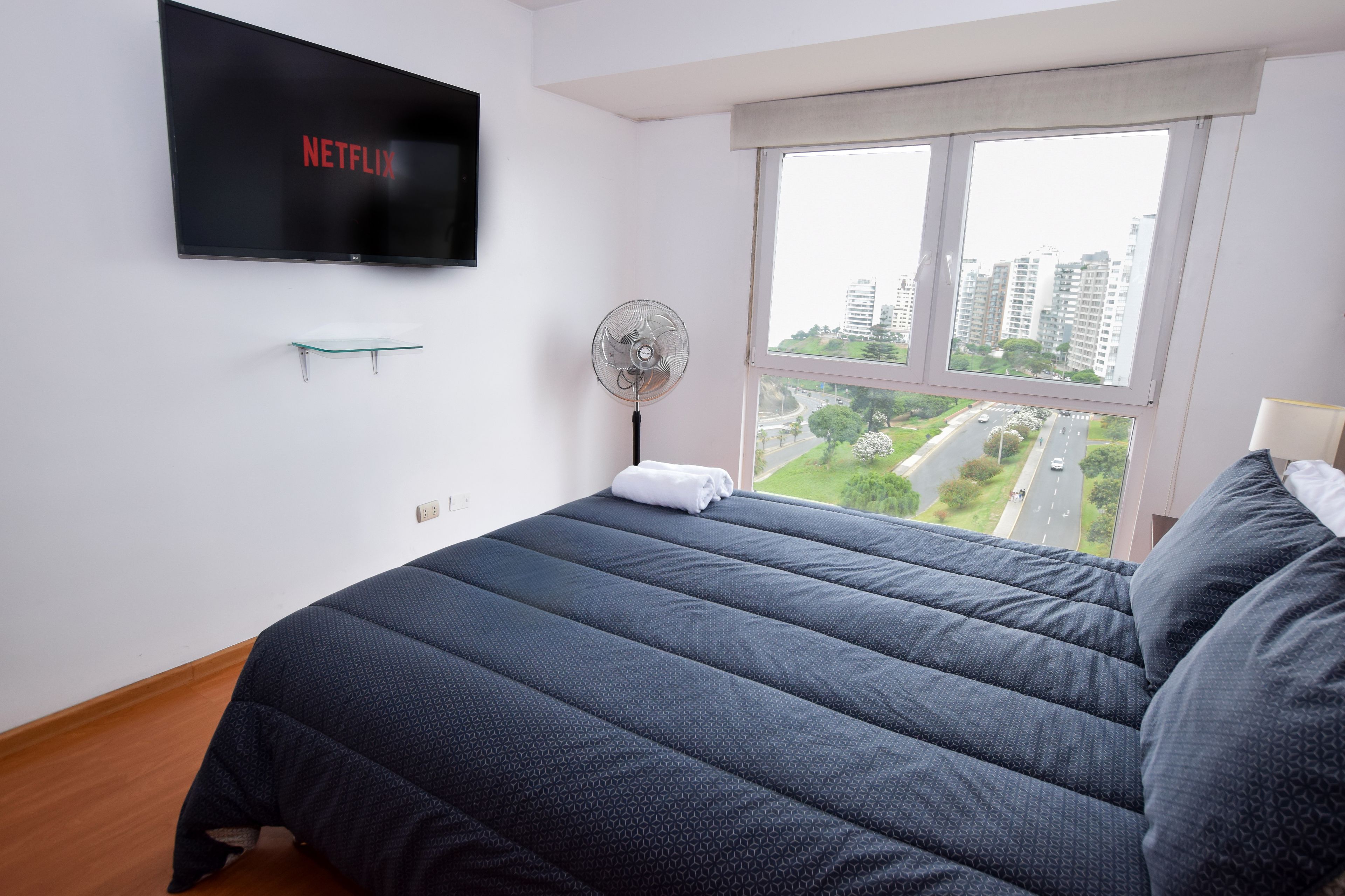Un dormitorio con cama, televisor montado en la pared, ventilador de pie y ventana con vistas a edificios y una carretera.