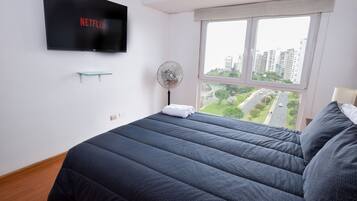 Un dormitorio con cama, televisor montado en la pared, ventilador de pie y ventana con vistas a edificios y una carretera.
