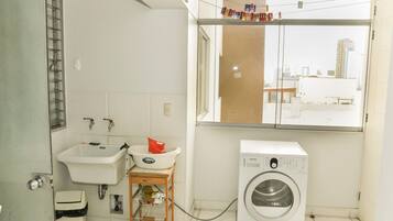 Pequeño lavadero con lavadora, lavabo y ventana que da a vistas de edificios.