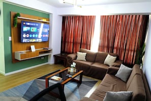 A living room with a sofa, a coffee table, a TV mounted on the wall, and a curtain. - Miraflores NEST Alcanfores, Near Kennedy Park, 2BR, AC, 1000 Mbps WiFi (Lima)
