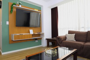 A living room with a flat-screen TV mounted on a wooden wall, a brown sofa, a glass coffee table, and a window with curtains. - Miraflores NEST Alcanfores, Near Kennedy Park, 2BR, AC, 1000 Mbps WiFi (Lima)