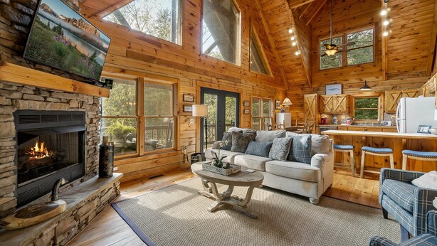 L’intérieur d’un chalet confortable, avec un foyer en pierre, un grand téléviseur fixé au mur, un coin cuisine et un espace salon comprenant un canapé et des fauteuils.