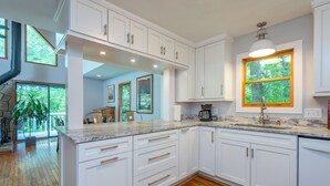 A modern kitchen with white cabinetry, a marble countertop, and a large window with a view of greenery. - Garden Cottage 2.5 Miles to Downtown With Private, Wooded Surroundings (Asheville)