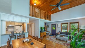 A spacious living area with a wooden ceiling, a dining table with chairs, a sofa, and a large window offering a view of greenery. - Garden Cottage 2.5 Miles to Downtown With Private, Wooded Surroundings (Asheville)