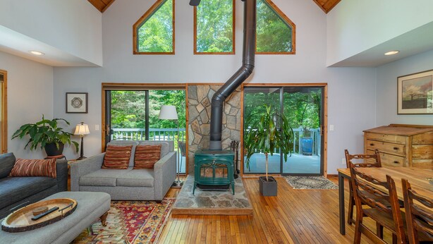 A spacious living room with a stone fireplace, wooden flooring, and large windows offering views of the outdoors. - Garden Cottage 2.5 Miles to Downtown With Private, Wooded Surroundings (Asheville)