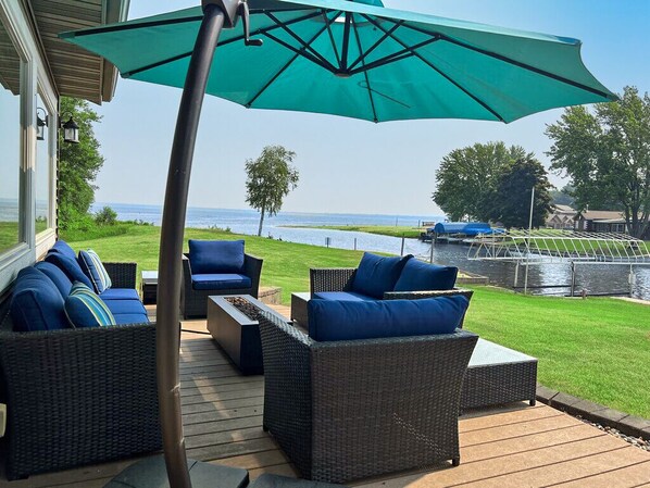 A deck with wicker furniture and a large umbrella, overlooking a body of water and a green landscape. - Lake Front House Directly On Castle Rock Lake - Dock, Beach & Near WI Dells (Mauston)