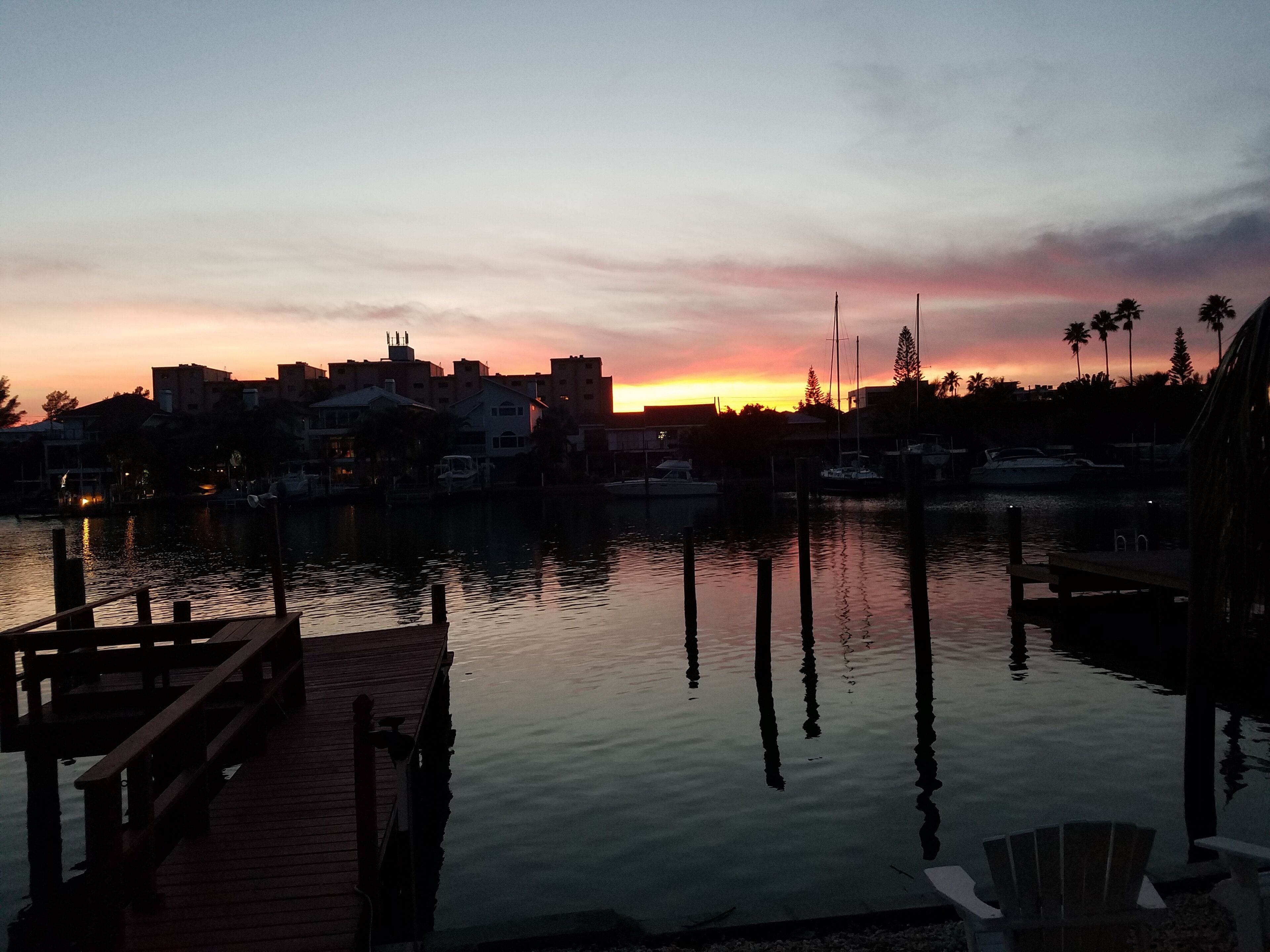 Un port de plaisance au coucher du soleil, avec une eau calme, des bateaux amarrés et des bâtiments en arrière-plan.