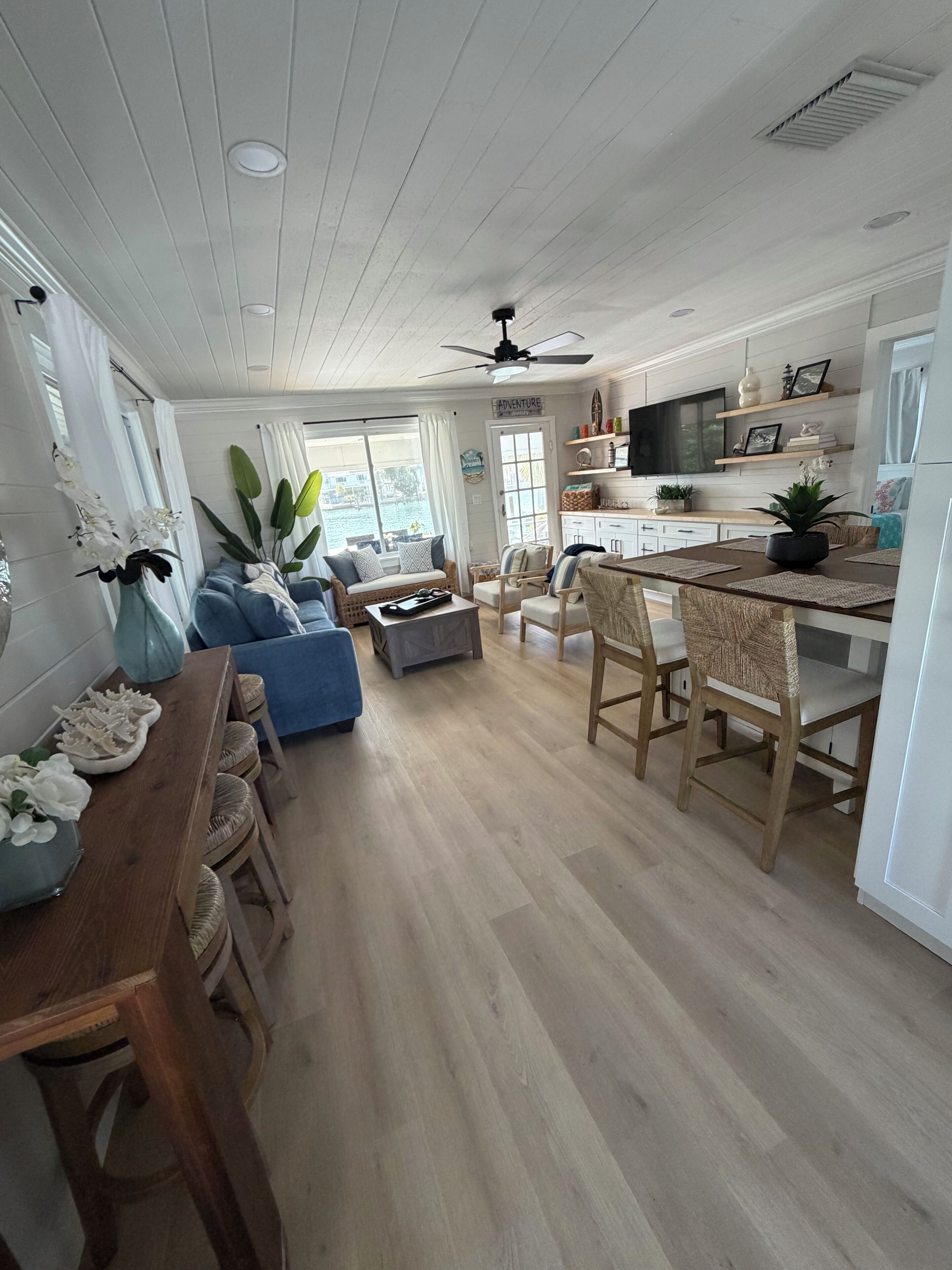 A spacious living room with a blue sofa, a wooden dining table, and a kitchen area with white cabinets and a TV.