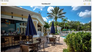 Un área de bar al aire libre con mobiliario de mimbre y sombrillas, una piscina y palmeras.