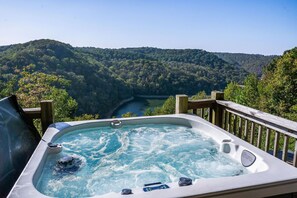 A hot tub with a wooden deck, surrounded by a forested landscape and a river.