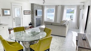 Ein modernes Wohnzimmer mit einem Glastisch, gelben Stühlen, einer Couch und Blick auf ein Gewässer durch große Fenster.