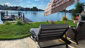 Eine Terrasse mit einer gemusterten Bank, einem Reetdachschirm und Blick auf einen See mit Häusern im Hintergrund.