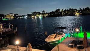 Eine Uferterrasse mit angedocktem Boot, Außenmöbeln und Blick auf das Wasser und den Stadtpanoramahimmel bei Nacht.