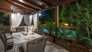 An outdoor dining area with a covered patio, wicker furniture, and a pool in the background. - Villa Eleonas (Zakynthos)
