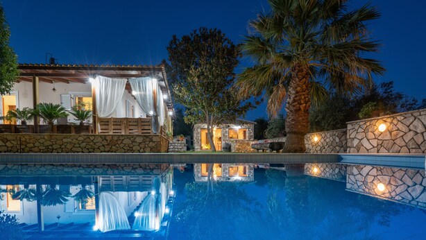 A poolside view of a resort with a stone wall, palm trees, and a building with a covered patio. - Villa Eleonas (Zakynthos)