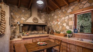 A rustic kitchen with a stone wall, a brick fireplace, wooden furniture, and a window overlooking greenery. - Villa Eleonas (Zakynthos)