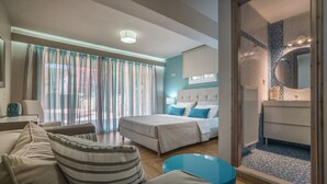 A modern hotel room with a bed, a sofa, a small table, and a bathroom with a tiled shower area. - Villa Eleonas (Zakynthos)