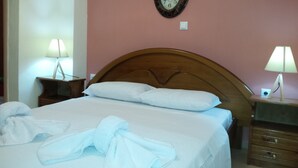 A neatly made bed with white linens and towels, a wooden headboard, a wall clock, and bedside lamps on wooden nightstands. - Rubini Home III 3 minutes from sea by feet (Chania)