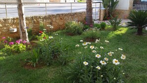 A garden with various flowers and plants, a stone wall, and a road in the background. - Rubini Home III 3 minutes from sea by feet (Chania)