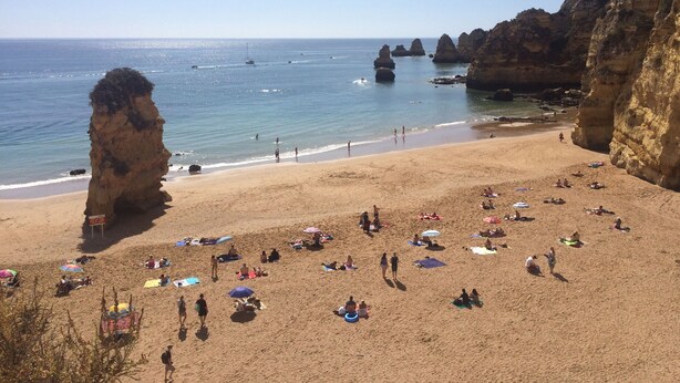 Une plage où des gens se prélassent au soleil et se baignent, entourée de falaises rocheuses et d’une eau d’un bleu limpide.