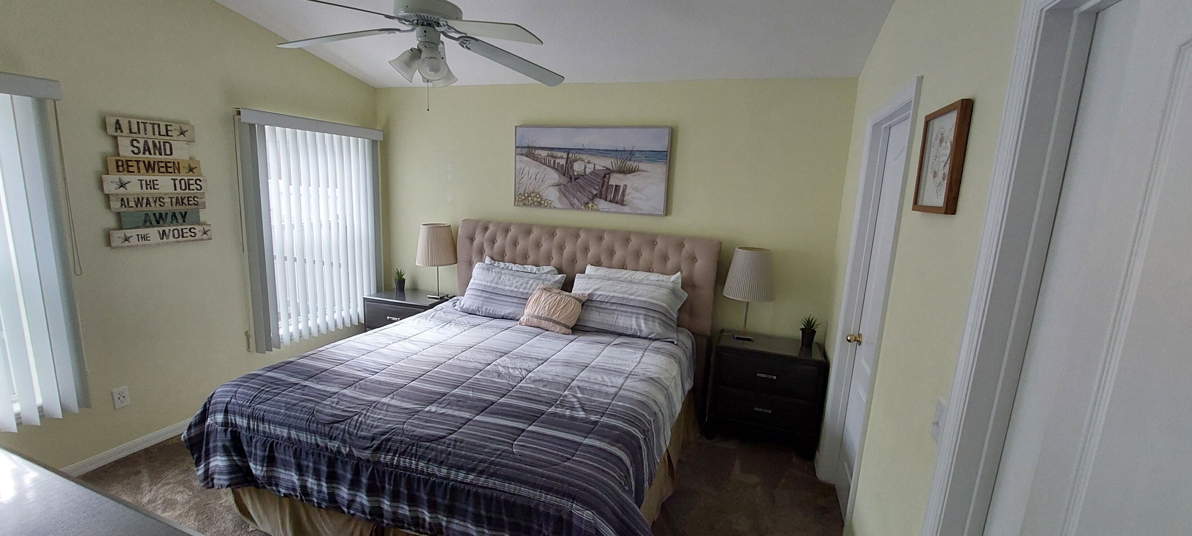 A bedroom with a bed, a ceiling fan, a nightstand, a lamp, and a wall with a beach-themed painting.