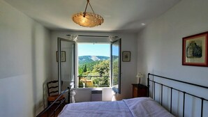 A bedroom with a bed, a chair, a bedside table, a hanging light fixture, and a large window with a view of greenery and mountains.