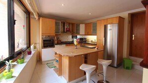A modern kitchen with wooden cabinets, a central island with stools, a stainless steel refrigerator, and a window with potted plants. - ViVa Royal (Arona)