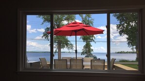 Blick aus einem Fenster auf einen See, mit einem roten Sonnenschirm und Sitzgelegenheiten im Freien.
