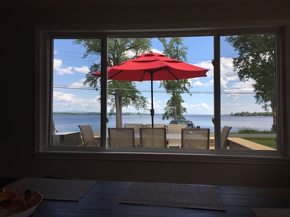 Une vue depuis une fenêtre donnant sur un lac, avec un parasol rouge et des sièges installés à l’extérieur.