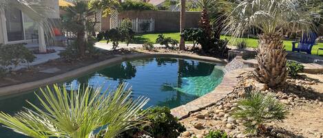 A swimming pool surrounded by palm trees and rocks, with a view of mountains in the background.