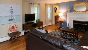 A living room with a leather sofa, a TV on a wooden stand, a fireplace, and a painting on the wall. - 4 Bedrooms, 2 Baths, Sleeps 8-10 (Chatham)