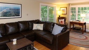 A living room with a brown leather sofa, a small wooden table, and a large window with a view of greenery. - 4 Bedrooms, 2 Baths, Sleeps 8-10 (Chatham)