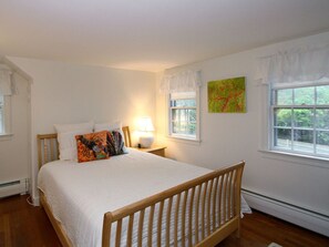 A bedroom with a wooden bed, white bedding, a bedside lamp, a window with a view of greenery, and a colorful abstract painting on the wall. - 4 Bedrooms, 2 Baths, Sleeps 8-10 (Chatham)