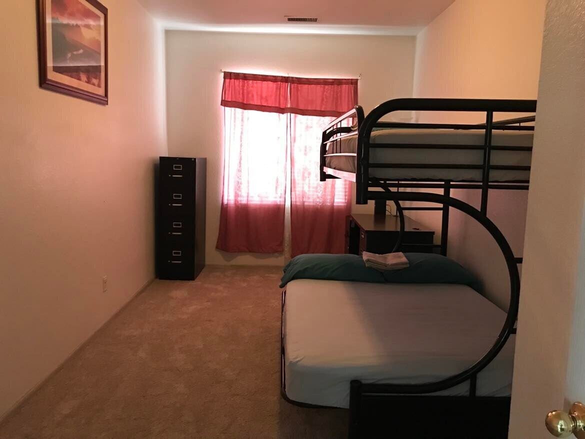 A room with bunk beds, a small cabinet, and a window with red curtains.