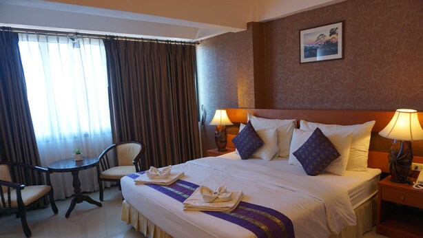 A hotel room with a large bed, two bedside tables with lamps, a small round table with chairs, and a window with curtains. - Kirimathani Hotel (Mueang Loei)