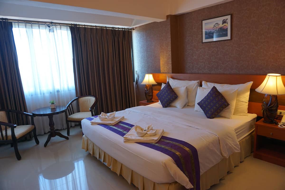 Habitación de hotel con una cama grande, dos mesitas de noche con lámparas, una mesa redonda con sillas y una ventana con cortinas.