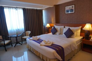 Quarto de hotel com uma cama grande, duas mesas de cabeceira com luminárias, uma mesinha redonda com cadeiras e uma janela com cortinas.