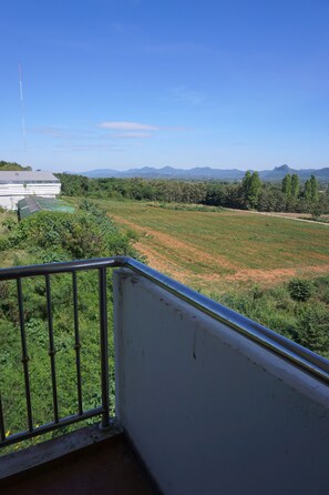 Uma varanda com grades de metal que dá para um campo gramado, um edifício com telhado verde e montanhas distantes sob um céu limpo.