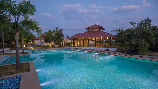A swimming pool with a covered area, surrounded by palm trees and a building with a red roof. - Tuli Veer Bagh Resort (Ramtek)