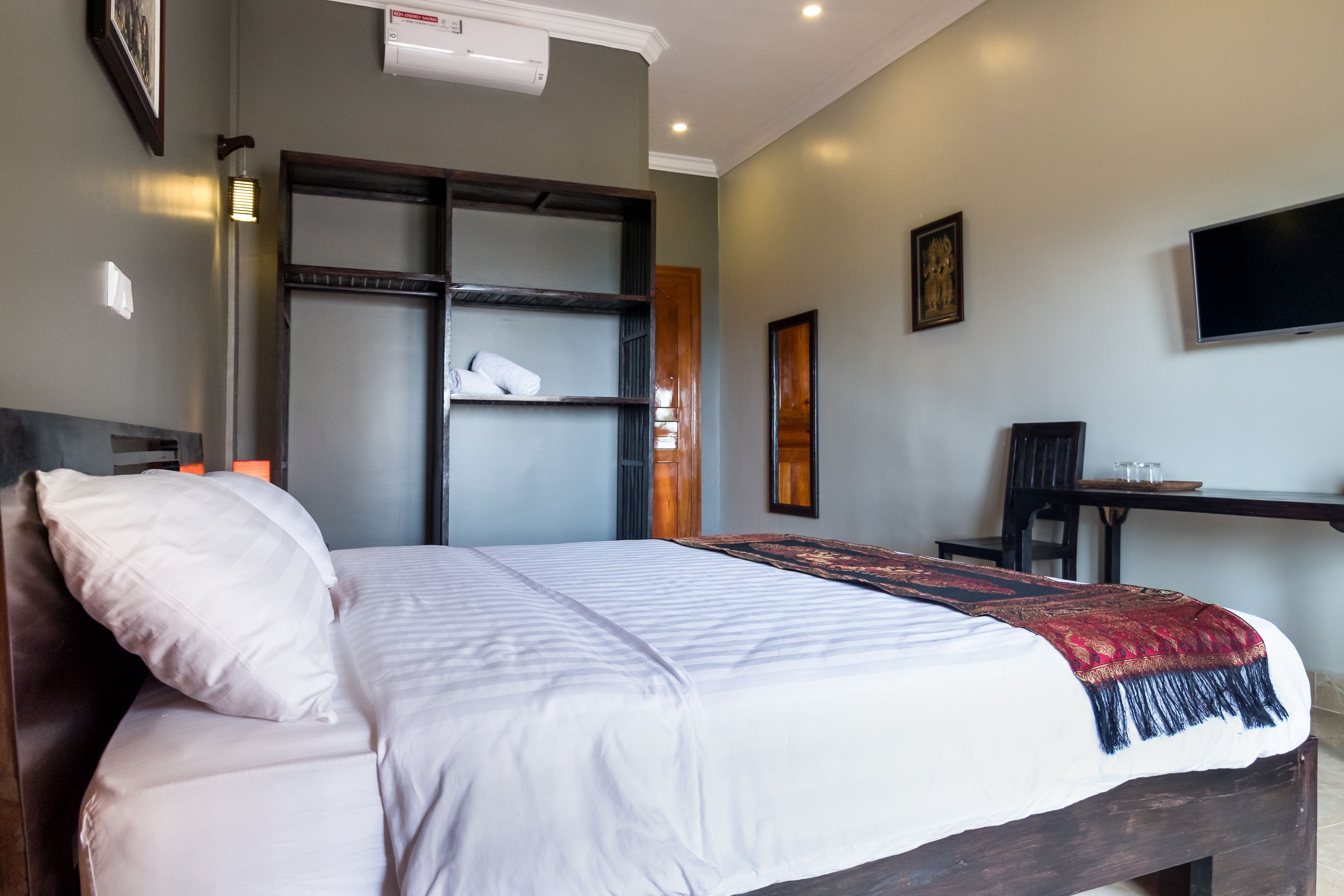 A neatly made bed with white linens and a red and black throw blanket. There is a wooden headboard, a wall-mounted shelf with folded towels, and a flat-screen TV on the wall.