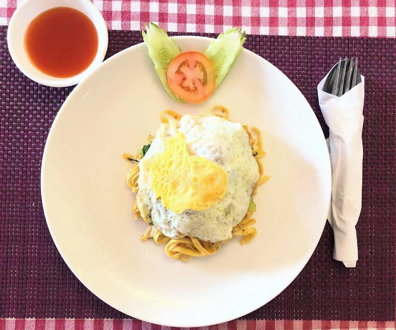 A plate of pasta with a fried egg on top, garnished with a slice of tomato and lettuce, accompanied by a small bowl of sauce, on a checkered tablecloth.