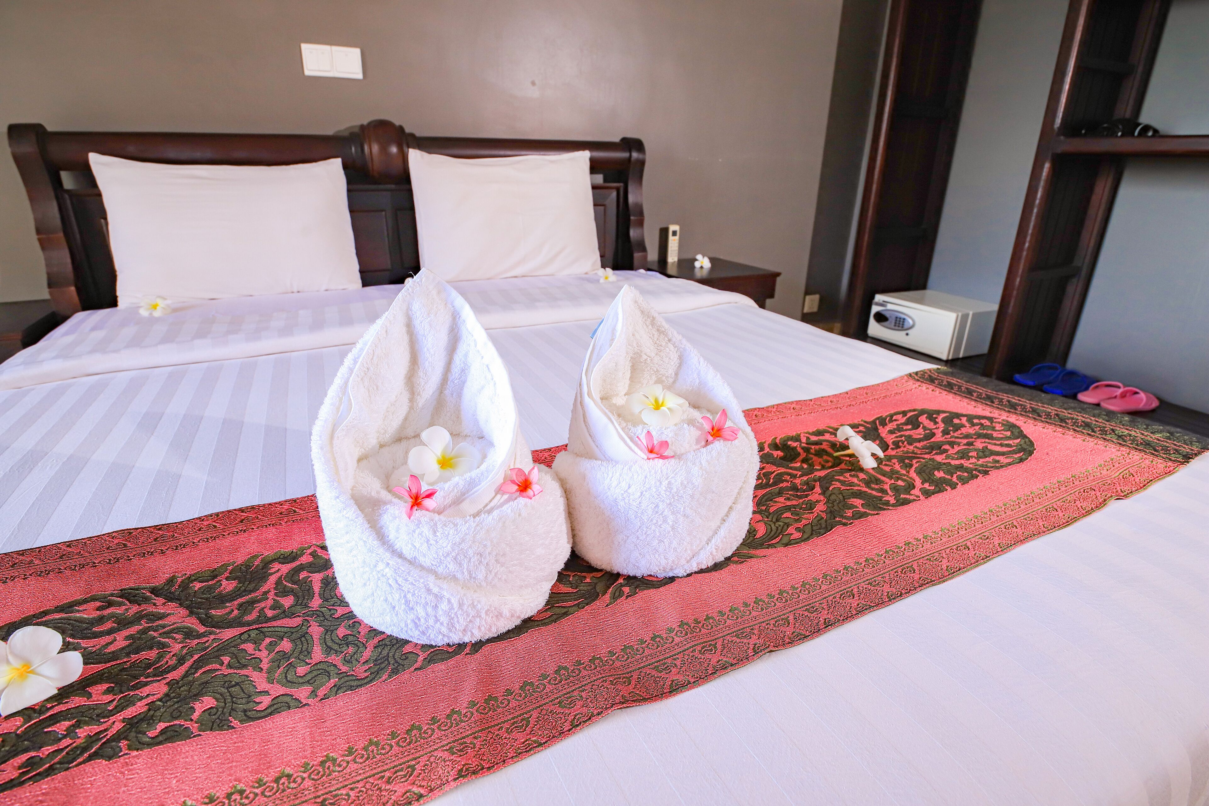 A neatly made bed with a red and black patterned runner, two towels with flowers, and a white flower on the bed.
